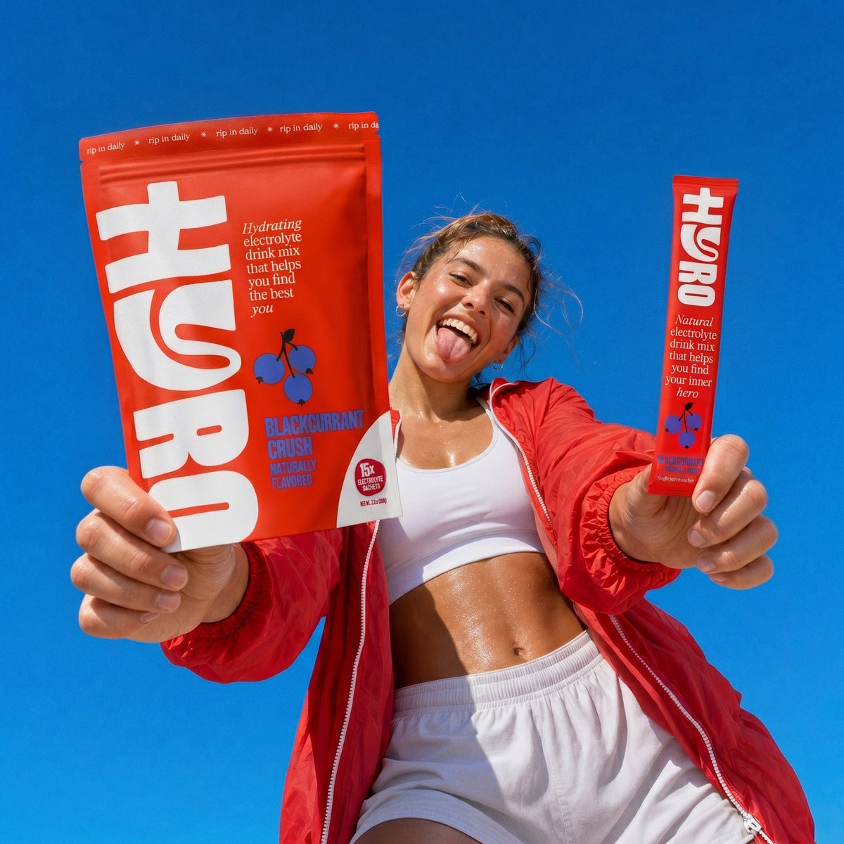Smiling young woman in red windbreaker holding a Hyro blackcurrant pouch and stick under a blue sky