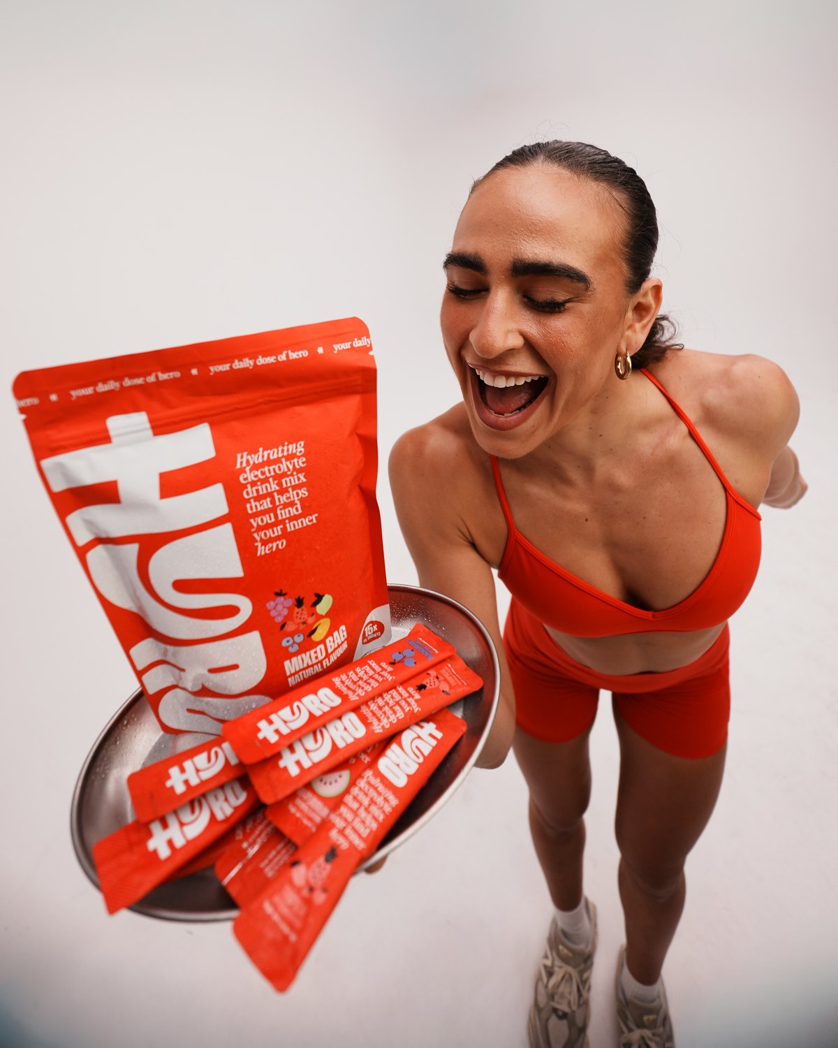 Laughing woman in red activewear holding a HYRO Mixed Bag pouch and a tray of HYRO sticks
